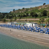 Spiaggia con file di ombrelloni blu, vicino a un tratto di strada e colline verdi.