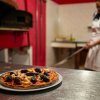 Pizza su un tavolo, forno a legna e pizzaiolo sullo sfondo.
