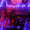 Persone sul palco con costumi e luci colorate in uno spettacolo teatrale.