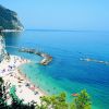 Spiaggia affollata con mare blu e scogliere in background.