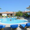 Piscina circolare con lettini blu, circondata da casette e palme sotto un cielo azzurro.