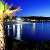 Una spiaggia illuminata di notte, con palme e il riflesso delle luci sull'acqua.