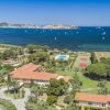 Un paesaggio costiero con edifici, piscina, campi da tennis e barche a vela in mare.