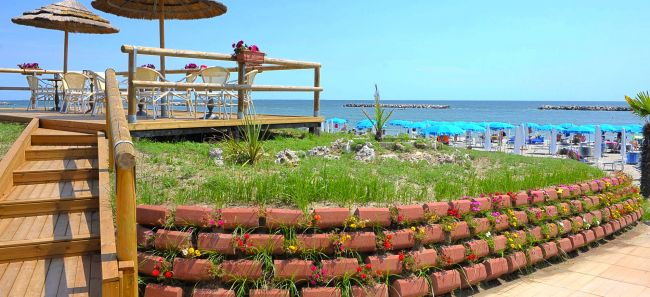 Un terrazzo con tavolini e sedie vista mare, ombrelloni di paglia e aiuole fiorite.