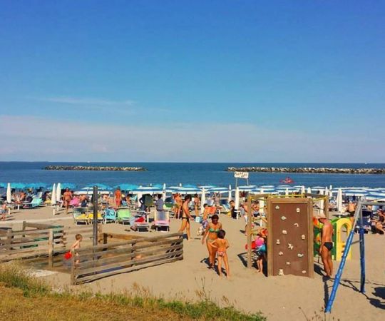 Spiaggia affollata, persone in costume, ombrelloni blu e giochi per bambini vicino al mare.