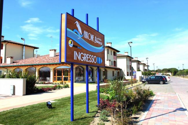 Cartello d'ingresso di un resort con edifici e auto parcheggiate.