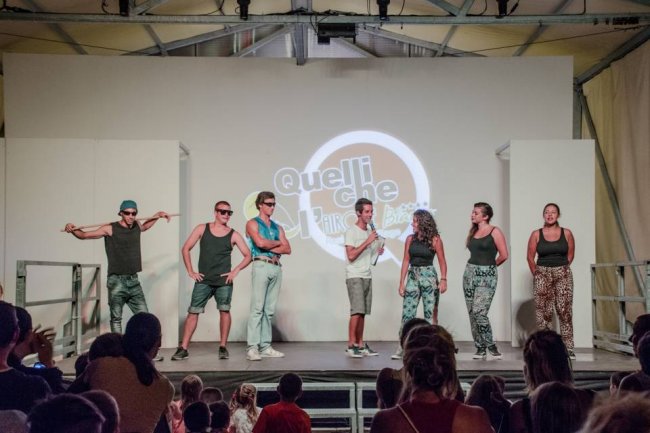 Sei persone su un palco partecipano a uno spettacolo con un pubblico seduto che li osserva.