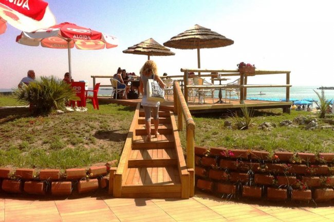 Una passerella in legno conduce a una terrazza con sedie e ombrelloni vista mare.