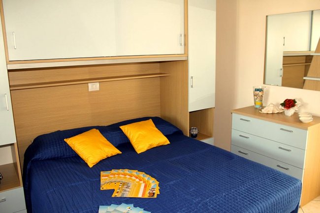 Camera con letto matrimoniale, cuscini gialli, mobili in legno chiaro, volantini sopra al letto.