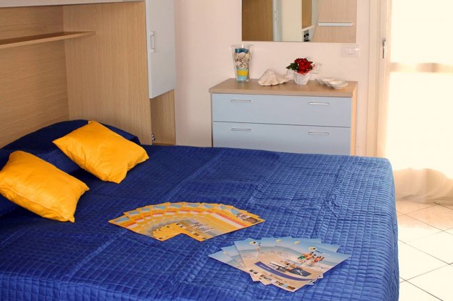 Camera da letto con copriletto blu, cuscini gialli e volantini sparsi su una superficie liscia.
