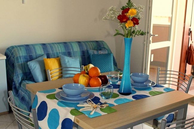 Tavolo apparecchiato con piatti blu, frutta, e vaso di fiori. Divano blu a quadri sullo sfondo.