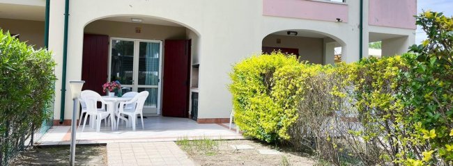 Patio esterno con tavolo bianco e sedie, circondato da siepi e piante verdi.