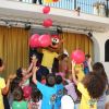 Bambini giocano con palloncini rossi intorno a una mascotte in un'area con tende gialle.