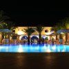 Piscina illuminata di notte, circondata da palme e edifici con arcate.