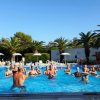 Persone fanno ginnastica in piscina sotto palme e ombrelloni bianchi.