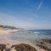 Una spiaggia rocciosa con mare calmo e ombrelloni in lontananza.