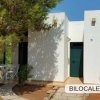 Bilocale bianco con porta verde e area esterna con tavolo e sedie, circondato da alberi.