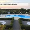Piscina all'aperto con sdraio e ombrelloni, circondata da siepi e vegetazione, vista mare.