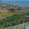 Vista dall'alto di una città costiera circondata da campi e alberi, con il mare sullo sfondo.