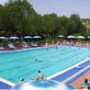 Una piscina affollata con lettini e ombrelloni blu, circondata da alberi verdi.