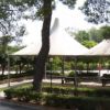 Un parco ombreggiato con pini, panchine e un'area coperta da tendoni.