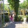 Bambini che camminano verso un parco giochi con altalene e uno scivolo giallo tra gli alberi.