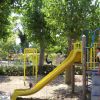 Un parco giochi con scivolo giallo e bambini che giocano, circondato da alberi.