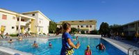 Piscina all'aperto con animatrice e persone in acqua. Edifici bianchi sullo sfondo.