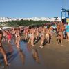 Spiaggia affollata con persone in costume e case bianche sullo sfondo.