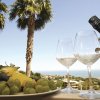 Vista su palme e mare con piatto di frutta, due bicchieri e una bottiglia di vino in primo piano.
