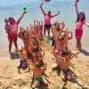 Bambini giocano in spiaggia con animatori che alzano le braccia e tengono palette colorate.