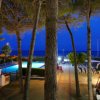 Vista notturna di una piscina illuminata, circondata da sedie e alberi con il mare sullo sfondo.