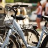 Bici parcheggiate all'aperto, alcune con cestini. Sfondo sfocato con persone.