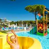 Bambino su uno scivolo giallo in un parco acquatico con piscina e alberi sullo sfondo.